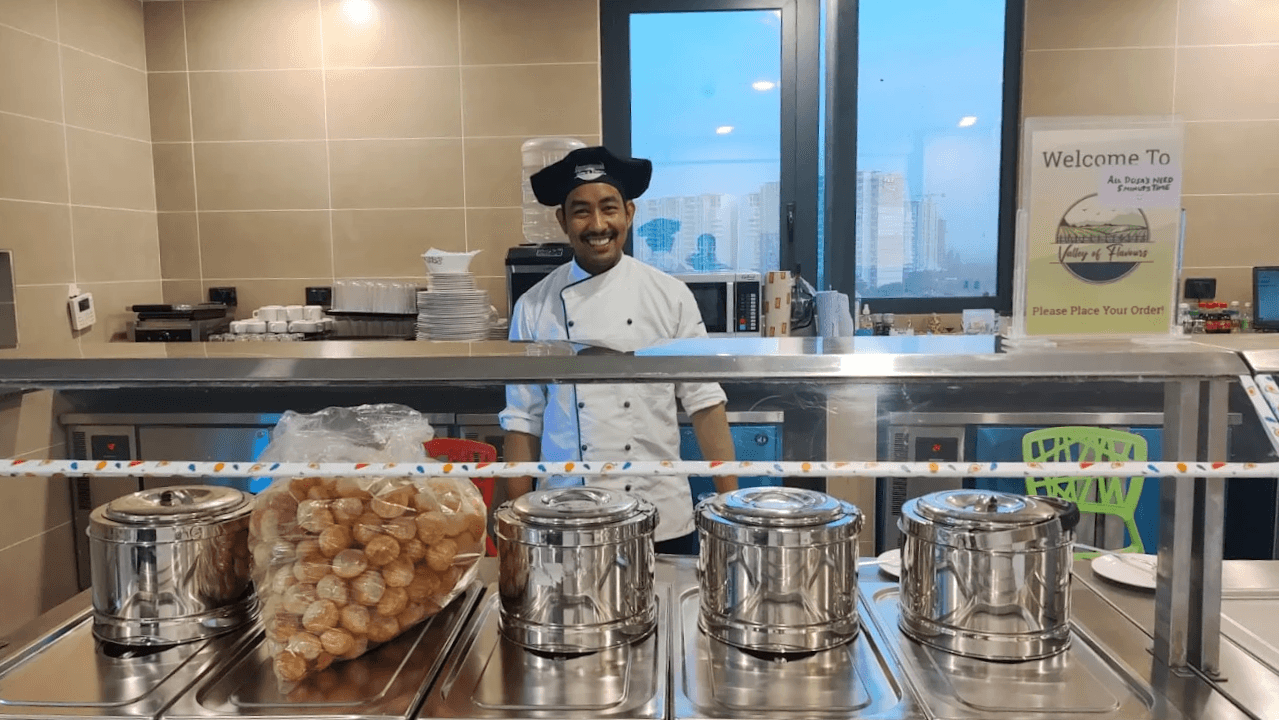 A chef in Valley of Flavours whites standing behind the service counter, with covered chafing dishes and a tray of golgappas in front of him.